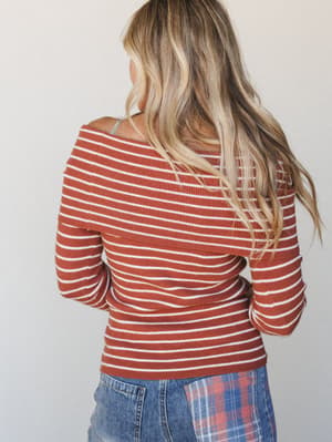 Shot from behind, a model in a rust-striped ribbed knit top with a wide off-shoulder neckline pairs it with plaid-patterned denim — relaxed, a little retro, and effortlessly wearable.