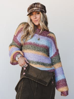 A relaxed, color-story knit sweater in earthy multicolor stripes styled with a plaid newsboy cap and dark corduroy skirt — the kind of outfit that feels like a farmers market Saturday done really, really right.