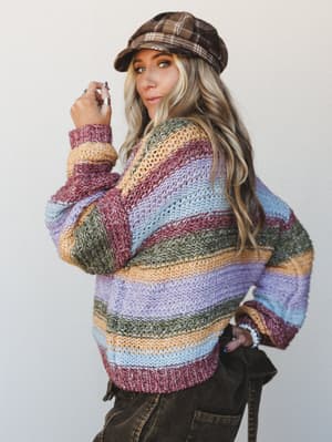 A model glances back over her shoulder wearing a chunky open-knit striped sweater in earthy multicolor stripes, paired with a plaid newsboy cap and dark corduroy pants — the whole look has that effortless, lived-in fall energy.