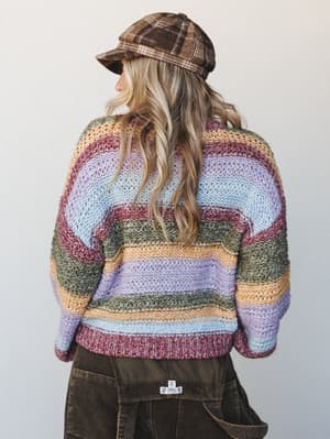 Shot from behind, a model in a chunky multicolor striped knit sweater, plaid baker boy cap, and dark olive corduroy pants — the kind of outfit that looks like it was pulled together on a perfect fall morning without overthinking it.