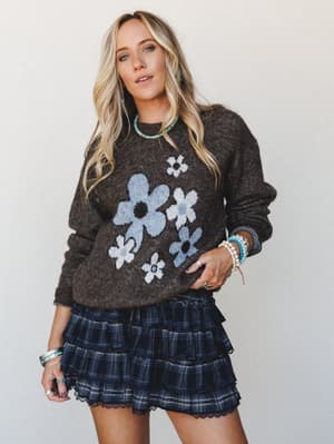 A relaxed, retro-leaning outfit pairing a charcoal knit sweater with oversized daisy intarsia with a tiered plaid mini skirt — styled with turquoise jewelry and stacked bracelets for that effortlessly collected, boho-vintage feel.