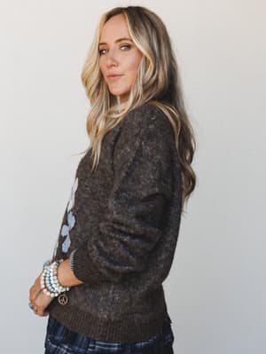 A model shot in three-quarter profile wearing a cozy dark brown knit sweater with a subtle floral motif, styled with plaid bottoms and stacked boho bracelets — the kind of outfit that feels like a warm drink on a cold afternoon.