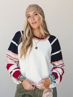 A relaxed, lived-in pullover top with color-blocked striped sleeves styled with a crochet beanie, stacked turquoise bracelets, and a delicate pendant necklace — the kind of outfit that looks effortless but clearly wasn't an accident.