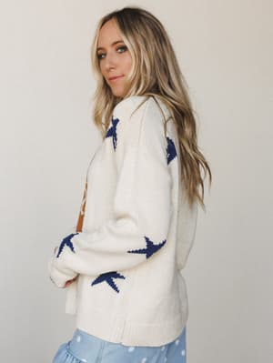 A cream chunky knit sweater with bold navy star intarsia details photographed from a three-quarter back angle — the kind of cozy piece that looks just as good on a coffee run as it does curled up on someone's couch.