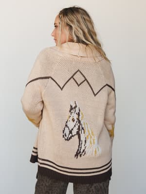 A back-view shot of an oversized knit cardigan in natural cream, featuring an intarsia horse graphic and mountain zigzag detail — the kind of sweater that makes you want to drive somewhere with a good view.