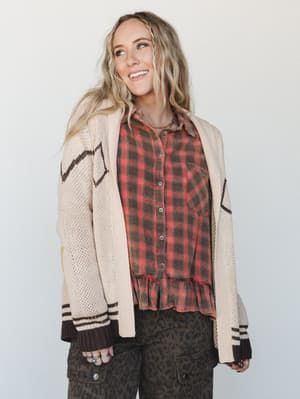 A layered fall outfit built around a cream knit cardigan with graphic intarsia detailing, worn open over a red plaid flannel with a ruffle hem, and paired with olive leopard print cargo pants — the kind of mix-and-match that looks effortless but is c