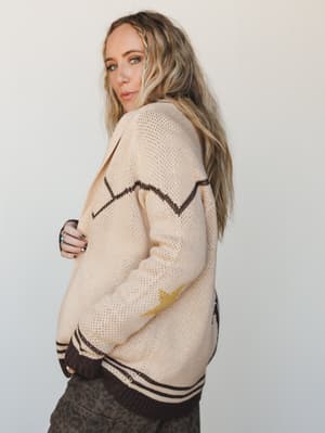 A relaxed, oversized knit cardigan in warm cream with dark brown zigzag intarsia, a gold lightning bolt detail on the sleeve, and varsity-style ribbed hem — styled over leopard print pants for a cool, lived-in boho-vintage vibe.