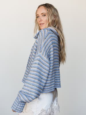 A model shot in three-quarter profile wearing a cozy striped knit sweater in soft blue and taupe tones — the kind of sweater that makes you want to find a window seat and stay there.