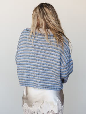 Shot from behind, a model wears a cropped knit sweater in soft periwinkle and taupe stripes paired with a white lace skirt — relaxed, effortless, and the kind of thing you'd reach for on a cool morning without thinking twice.