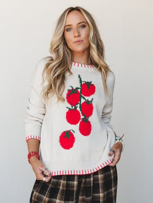 A cream knit sweater with a bold tomato vine intarsia graphic and red contrast stitching at the hem, cuffs, and neckline, styled with a brown plaid midi skirt and layered bracelets — the kind of piece that makes grocery runs feel like a personality s