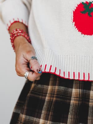 A close-up detail shot showing the ribbed hem of a cream knit sweater with a tomato intarsia print and red blanket-stitch trim, layered over a brown plaid skirt — the model's hand pulls the hem slightly, showing off stacked bracelets and a mix of rin