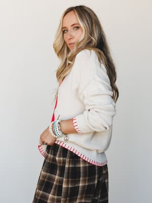 A cream knit sweater with red contrast stitching along the hem and cuffs is layered over a red top and paired with a dark plaid midi skirt — the kind of outfit that looks like it came together effortlessly but absolutely did not.
