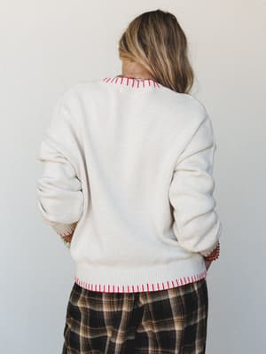 Shot from behind, a cream knit sweater with bright red whipstitch trim at the neckline, cuffs, and hem is paired with a dark plaid wide-leg pant — relaxed, a little retro, and very much the kind of outfit you wear when you want to look effortless but