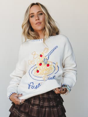 A cream knit sweater with a playful pasta bowl graphic and embroidered tomato details paired with a brown plaid tiered skirt — cozy and personality-forward, like something you'd wear to brunch and get complimented on all day.