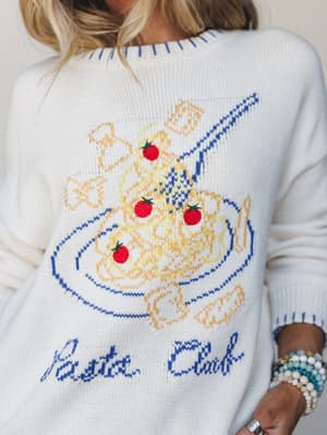 A close-up of a cream knit sweater with a playful 'Pasta Club' graphic — a bowl of spaghetti with cherry tomatoes rendered in blue and gold intarsia knit — worn with stacked beaded bracelets that feel like the whole outfit has a personality.