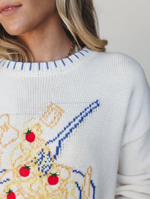 A close-up shot of an ivory knit sweater with a whimsical intarsia pasta bowl graphic — tomatoes, noodles, and all — worn with a delicate gold necklace and that quiet smile of someone who's already planning dinner.