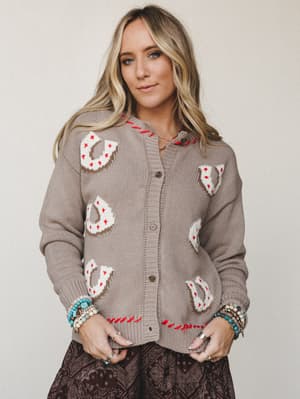 A cozy knit cardigan in warm greige with intarsia horseshoe motifs and red whipstitch trim, styled with a dark paisley skirt and stacked beaded bracelets — the kind of piece that makes a country music fan and a boho girl equally happy.