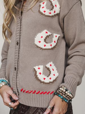 A close-up of a taupe knit cardigan covered in cream horseshoe intarsia motifs with red star embroidery accents and a red whipstitch hem — styled with stacked beaded bracelets and rings for a Western-boho feel that's equal parts cozy and characterful