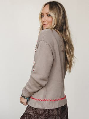 A model glances back over her shoulder showing off the back of a taupe knit cardigan with embroidered detailing and a bold red whipstitch hem, layered over a dark paisley skirt — the kind of outfit that looks like it came together effortlessly but ab