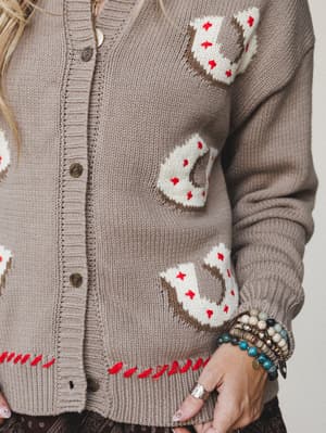 A close-up of a taupe knit cardigan covered in intarsia horseshoe motifs in cream and brown with red star accents, styled with stacked boho bracelets and a silver ring — the kind of piece that makes a whole personality statement without saying a word