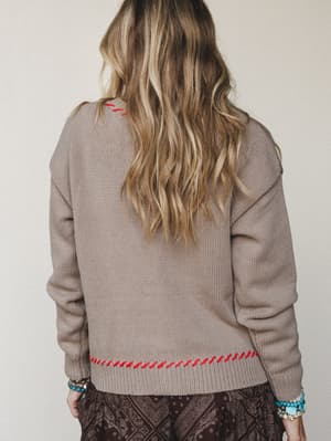 A back-view shot of a chunky knit sweater in warm greige, detailed with red whipstitch trim along the hem and neckline — layered over a dark paisley skirt with stacked turquoise and beaded bracelets adding that easy boho finishing touch.