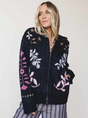 A flowy-haired blonde model gazes off to the side in a chunky navy zip-up sweater cardigan covered in intarsia floral and botanical motifs — the kind of piece that makes a Tuesday feel intentional.