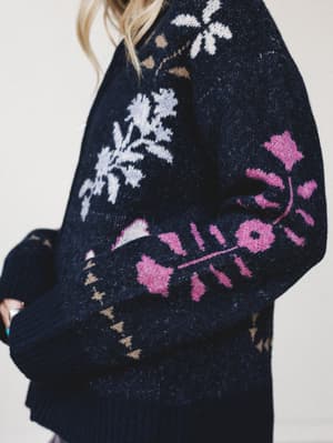 A close-up of an oversized navy knit sweater with a bold intarsia floral pattern — hot pink blooms and white botanical motifs give it that cozy-but-expressive energy perfect for a fall day that deserves more than a plain pullover.