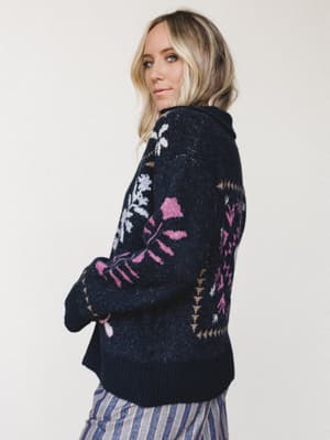 A blonde model looks back over her shoulder in a cozy navy intarsia knit sweater with floral and geometric motifs in pink, white, and tan — the kind of sweater you reach for on a crisp fall morning without thinking twice.