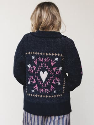 Shot from behind, a model wears a navy knit sweater with a bold intarsia heart-and-floral motif on the back, paired with striped wide-leg pants — cozy and a little romantic, like something you'd reach for every single weekend from October through Feb