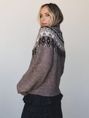 A model glances back over her shoulder wearing a cozy Fair Isle turtleneck sweater in heather mocha, the yoke pattern doing all the talking against a soft neutral studio backdrop — paired with a peek of a dark floral skirt below.