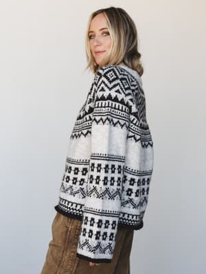 A side-profile shot of a cozy Fair Isle knit sweater in cream and black with geometric and floral motifs, paired with olive wide-leg pants — the kind of sweater that earns compliments at the farmer's market and stays in rotation all season.