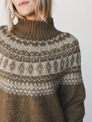 A close-up model shot of a cozy Fair Isle turtleneck sweater in earthy olive brown, with a detailed yoke pattern in cream and warm brown tones — the kind of sweater that makes you want to plan a weekend somewhere with fireplaces.