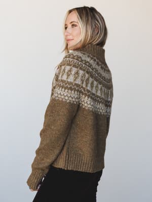 A cozy Fair Isle turtleneck sweater in heathered olive brown, shot from a three-quarter back angle — the kind of sweater that makes you want to find a window seat and a warm drink.