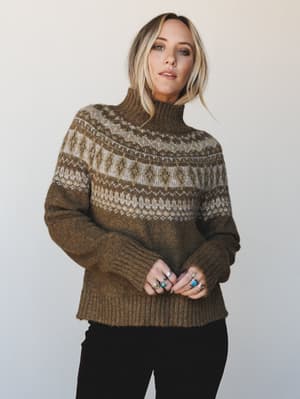 A cozy Fair Isle turtleneck sweater in warm olive brown layered over black pants, styled with a stack of silver and turquoise rings — the kind of sweater that makes a Tuesday feel intentional.