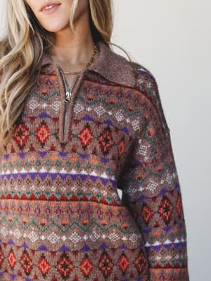 A close-up of a multicolor fair isle pullover sweater with a half-zip collar, worn by a blonde model against a soft neutral background — cozy, earthy, and the kind of thing you'd reach for every single weekend.
