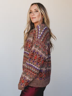 A model shot in three-quarter profile wearing an oversized Fair Isle knit sweater in warm brown with pops of rust, purple, and teal — the kind of sweater that makes you want to find a window seat and a hot drink.