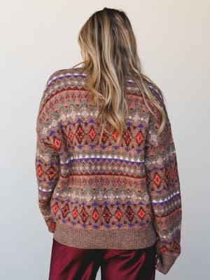 A back-view shot of a cozy Fair Isle knit sweater in warm brown with multicolor geometric patterning, paired with deep burgundy satin pants — the kind of sweater that makes a Saturday feel intentional.