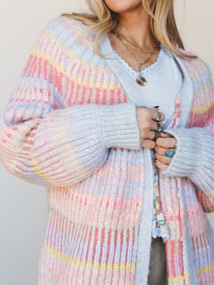 A chunky ribbed-knit cardigan in a dreamy pastel multi stripe worn open over a light blue lace-trim tank, styled with layered gold necklaces and a stack of silver and turquoise rings — the kind of sweater you reach for when you want to feel soft and