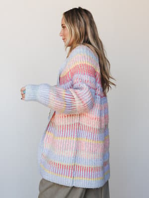 A long, chunky-knit cardigan in a dreamy mix of pastel stripes — lavender, blush pink, coral red, and soft yellow — worn side-profile over olive pants, with a relaxed, cozy feel that's equal parts nostalgic and wearable.