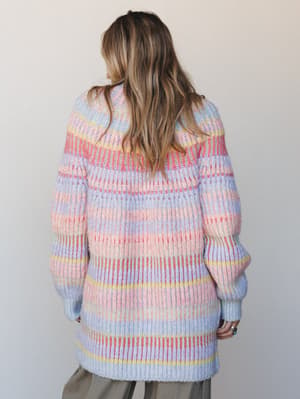 Shot from the back, a model wears an oversized striped knit cardigan in soft pastel stripes — lavender, blush, coral, and butter yellow layered together in a cozy, color-happy way that feels like spring in sweater form.