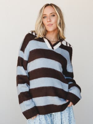 An oversized striped knit sweater in bold chocolate brown and powder blue, styled with a floral skirt peeking out at the hem — the kind of effortless layering that looks like it happened on purpose.