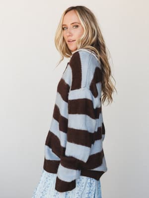 A side-profile shot of a model in an oversized striped knit sweater — bold chocolate brown and powder blue stripes with a soft, fuzzy texture — layered over a delicate floral midi skirt, shot against a clean light studio backdrop.