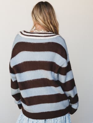 Shot from behind, a model layers an oversized striped knit sweater in powder blue and deep chocolate brown over a floral skirt — the kind of cozy, collected combo that looks like it came together effortlessly.