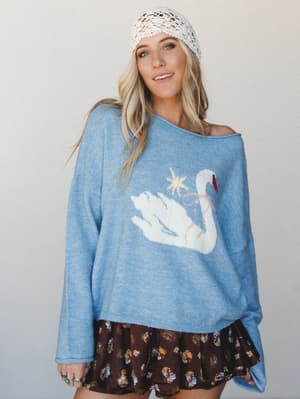 A breezy, whimsical sweater moment — periwinkle blue knit with a swan graphic worn off-shoulder, layered over a dark floral mini skirt and topped with a cream crochet beanie for that effortlessly collected, storybook-cozy look.