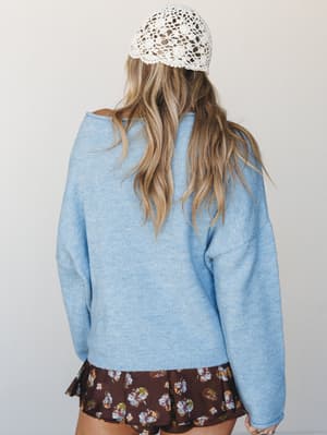 Shot from behind, she's wearing a soft sky blue oversized sweater layered over a dark floral mini skirt, topped with a cream crochet beanie — the kind of effortless mix that looks accidental but really isn't.