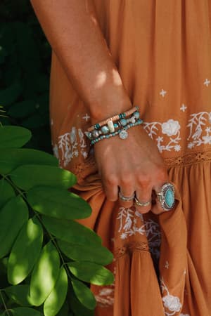 A person wearing a rust-orange dress with white embroidery holds the hem, displaying silver rings and the San Antonio Bracelet Set - Turquoise. Green leaves appear on the left side of the image.