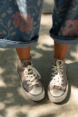 Person wearing blue floral pants and Chantel Studded High Top Sneaker in Mocha, featuring rhinestone detail, white laces, and studded high-top soles, standing on a sunlit concrete surface.