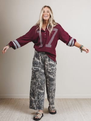 Woman wearing a burgundy oversized shirt with number 17 and camouflage pants against a plain background