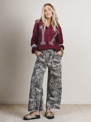 Woman wearing a burgundy sweatshirt with a number and camouflage pants indoors.