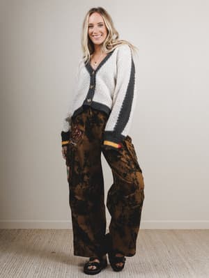 Woman wearing a white and black cardigan with brown patterned pants against a plain background